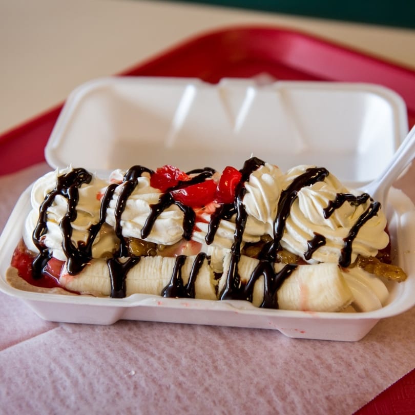 A banana split with whipped cream, cherries, and chocolate syrup in a white takeout container.