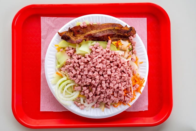 A large salad with ham, bacon, onions, and carrots on a white plate and red tray.