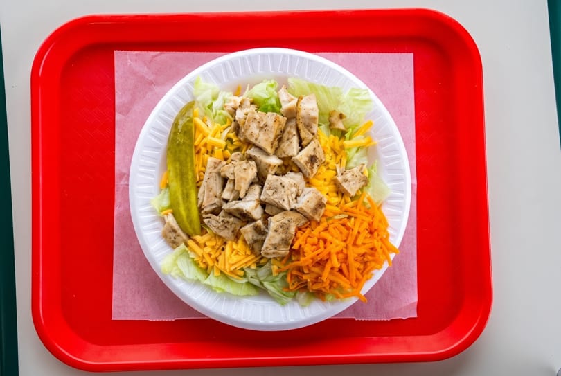 A chicken salad with lettuce, shredded cheese, carrots, and a pickle spear on a red tray.