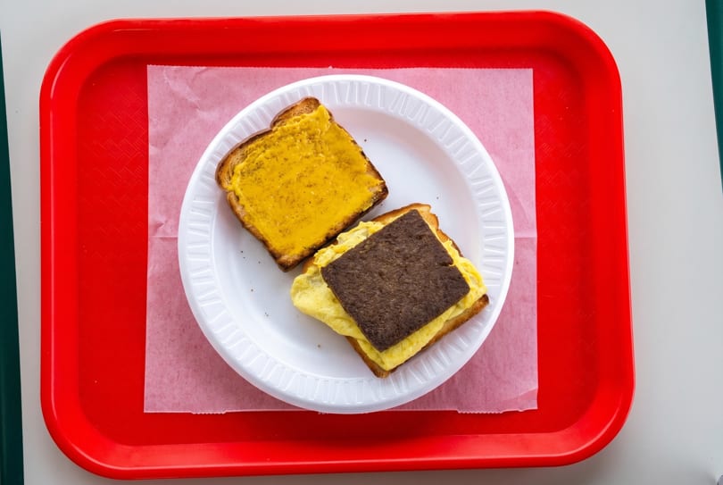 Two pieces of toast on a white paper plate, one with cheese, the other with egg and sausage.